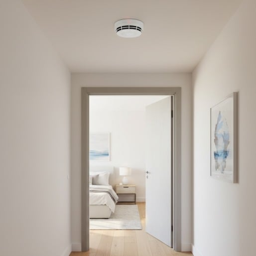 A white smoke detector mounted on the ceiling of a hallway looking into a bedroom.